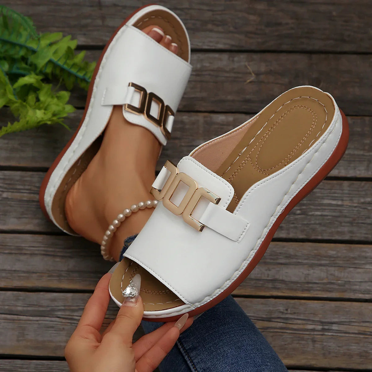 White slide sandals with gold buckle, minimalist design, worn on wooden deck with greenery