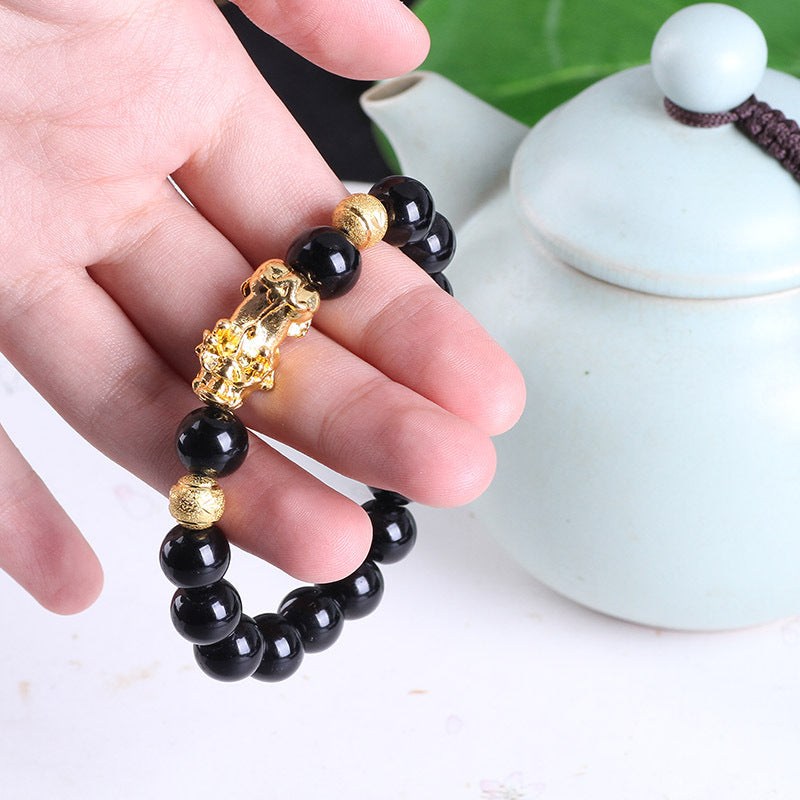 Black onyx beaded bracelet with gold Pi Xiu charm, held in hand beside ceramic teapot