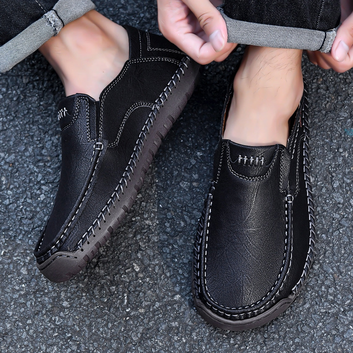 Black leather slip-on loafers with contrast stitching, worn with cuffed jeans on asphalt