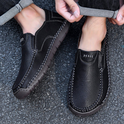 Black leather slip-on loafers with contrast stitching, worn with cuffed jeans on asphalt