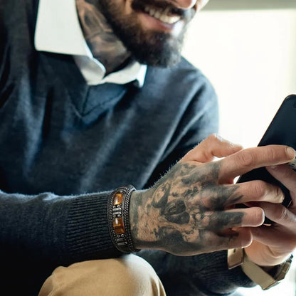 Man with tattooed hand wearing amber and metal bracelet, using smartphone