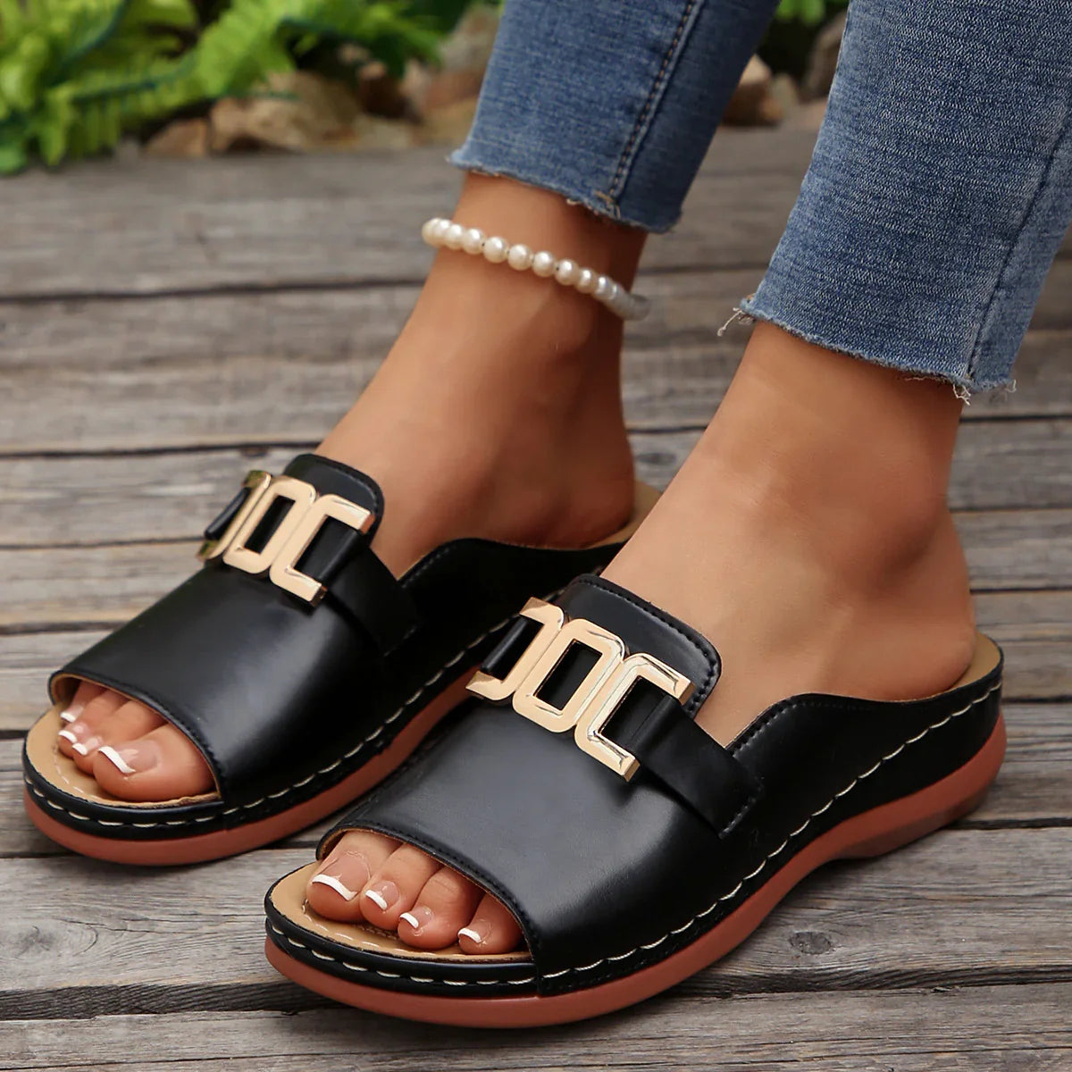 Black leather slide sandals with gold buckle detail, worn with jeans on wooden deck