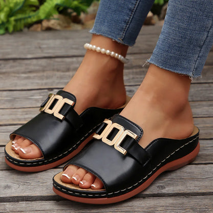 Black leather slide sandals with gold buckle detail, worn with jeans on wooden deck
