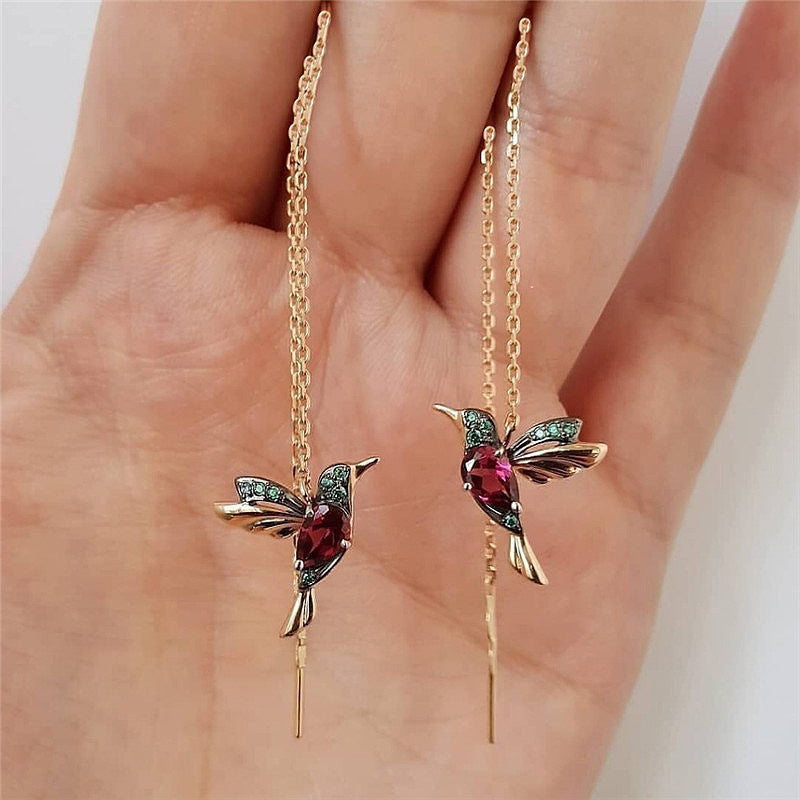 Gold hummingbird earrings with red gemstone bodies and green wing accents, held in hand.